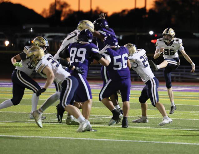 People in football uniforms are playing on a field at sunset with one person kicking the ball and others preparing to block or tackle. Description generated by AI