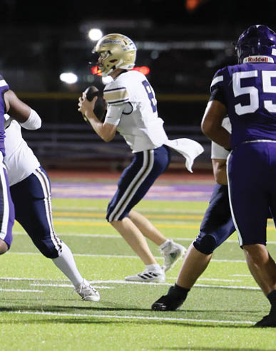 A person in a white football uniform is preparing to throw a football while being protected by teammates in purple uniforms on a field at sunset. Description generated by AI