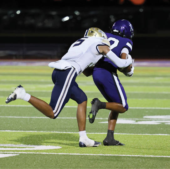 Two people in football uniforms are engaged in a tackle on a field under stadium lights during a game. Description generated by AI