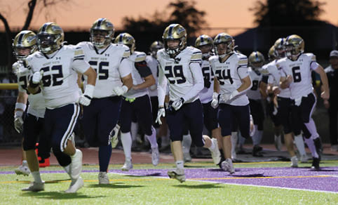 A group of people wearing football uniforms are running onto a field at dusk with trees and lights in the background. Description generated by AI