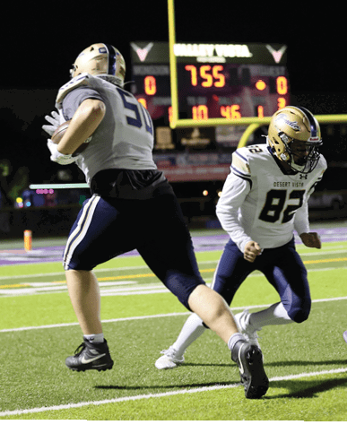 Two people in football uniforms are actively playing on a field at night with a scoreboard visible in the background. Description generated by AI