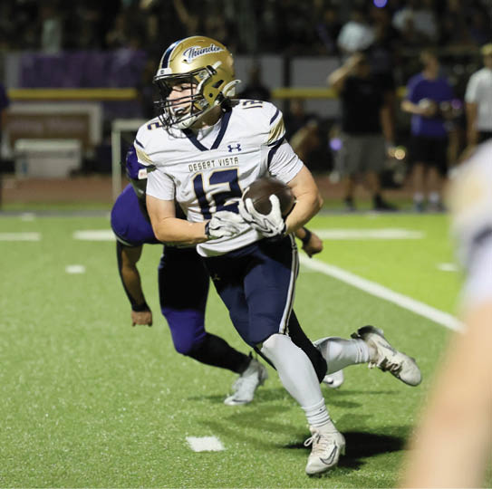 A person wearing a white and navy football uniform with a gold helmet is running on a football field while holding a football, with another person in a dark uniform chasing behind. Description generated by AI