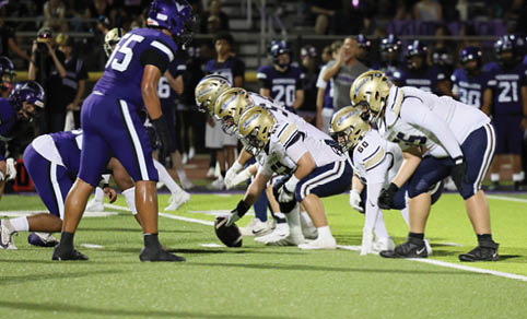 Two opposing football teams are lined up at the line of scrimmage on a field under stadium lights, with one team in white and gold uniforms and the other in purple uniforms. Description generated by AI