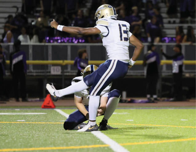 A person in a white sports uniform is kicking a football held by another person kneeling on a green field with spectators in the background. Description generated by AI