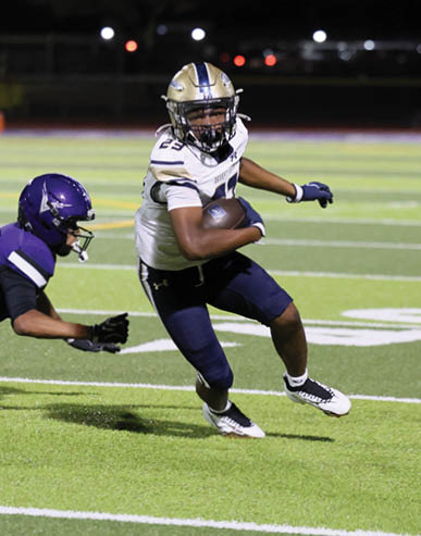 A person wearing a white and blue football uniform is running with the ball while another person in a purple uniform reaches out to try to tackle them on a football field at night. Description generated by AI