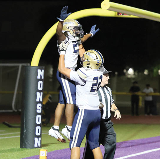 Two people in football uniforms celebrate near the end zone on a purple field with a referee standing nearby. Description generated by AI