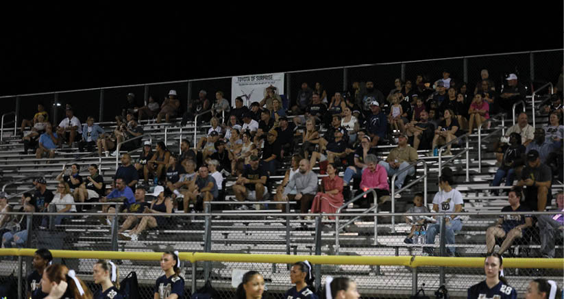 A crowd of people is seated on bleachers at night watching an event while several people in sports uniforms stand in front of the bleachers. Description generated by AI