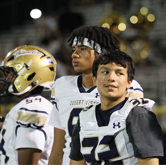 Three people wearing football uniforms stand together on a field with blurred stadium seating and lights in the background. Description generated by AI