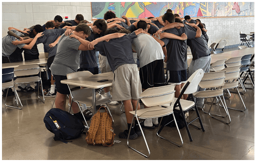 A group of people wearing casual clothing are standing closely together near a row of white folding chairs in an indoor setting with a polished concrete floor. Description generated by AI