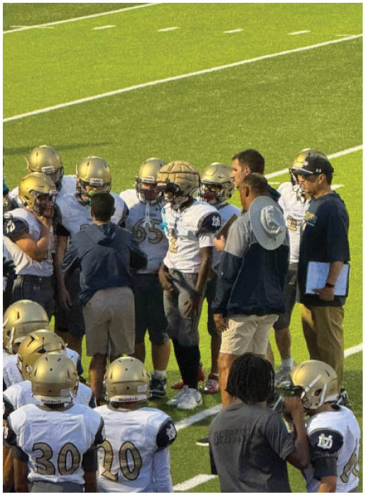 A group of football players wearing helmets and uniforms are gathered on a field with two coaches giving instructions. Description generated by AI