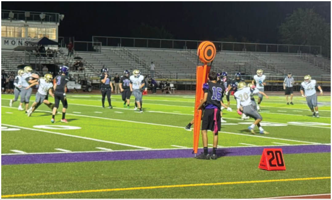 People in sports uniforms are playing football on a field at night with empty bleachers in the background. Description generated by AI