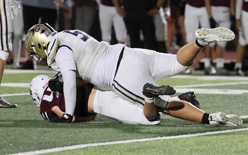 Two people in football uniforms are on the field with one tackling the other while other players and a coach stand in the background. Description generated by AI