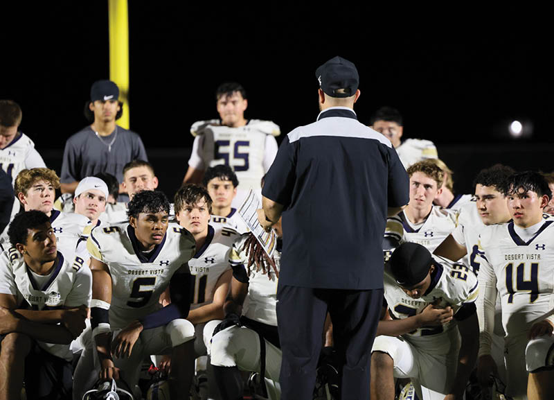 A group of people wearing football uniforms are gathered around a person in a coach's outfit who is speaking to them on a field at night. Description generated by AI