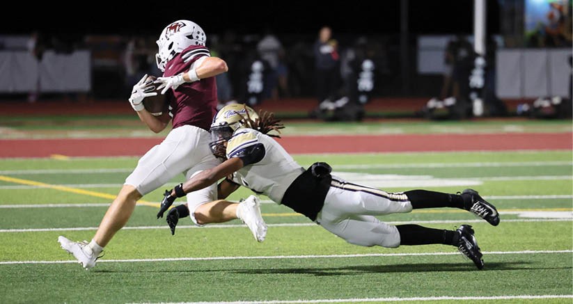 A person in a maroon jersey and white pants is running with a football while another person in a white jersey and black pants is diving to tackle them on a football field at night. Description generated by AI