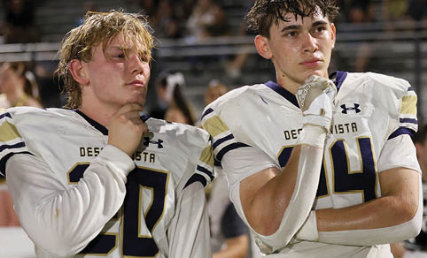 Two people in football uniforms stand side by side on a field, both resting their chins on their hands with thoughtful expressions. Description generated by AI