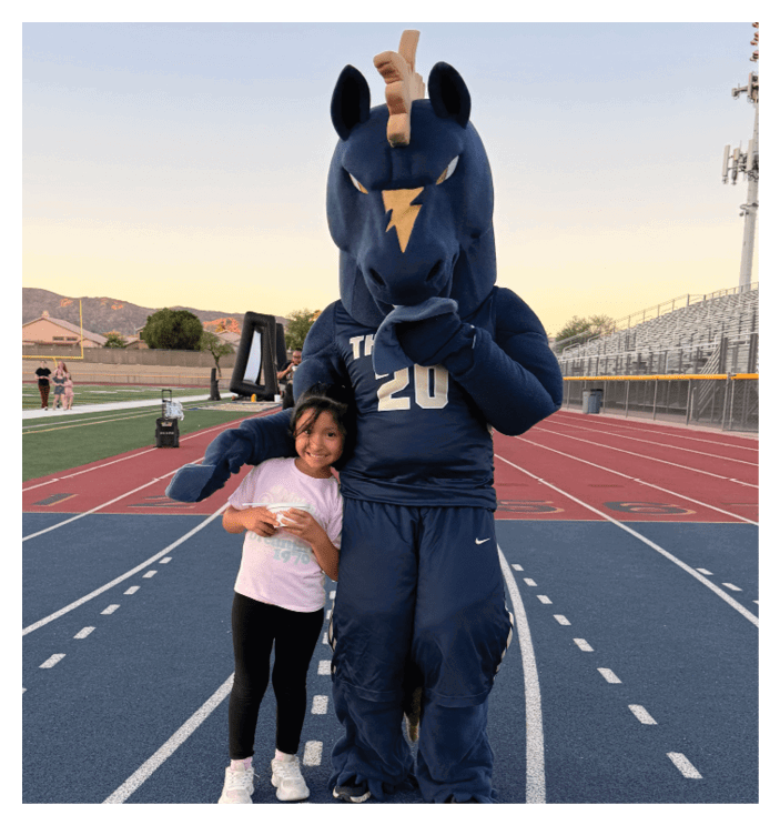 A person in a blue mascot costume is standing next to a child wearing a white shirt and black pants on a blue running track. Description generated by AI