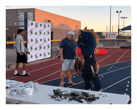 Two people wearing casual clothing and hats stand near a table with scattered items on a running track at sunset. Description generated by AI