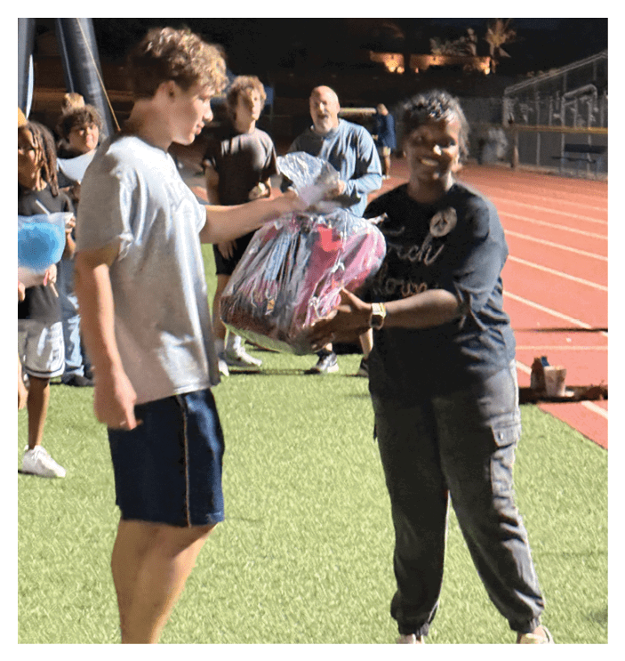 Two people stand on a sports field near a running track, with one person holding a wrapped gift while the other reaches out toward it. Description generated by AI