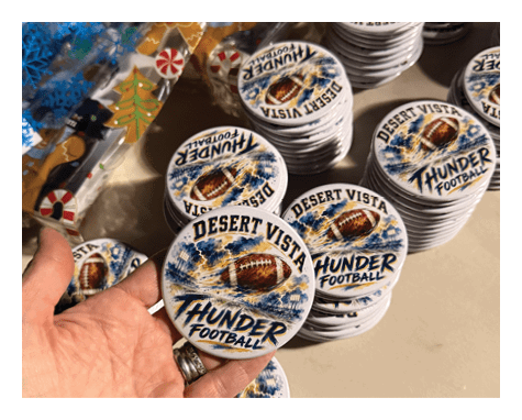A hand is holding a round button with a football design and the text “Desert Vista Thunder Football" among many similar buttons stacked on a table. Description generated by AI