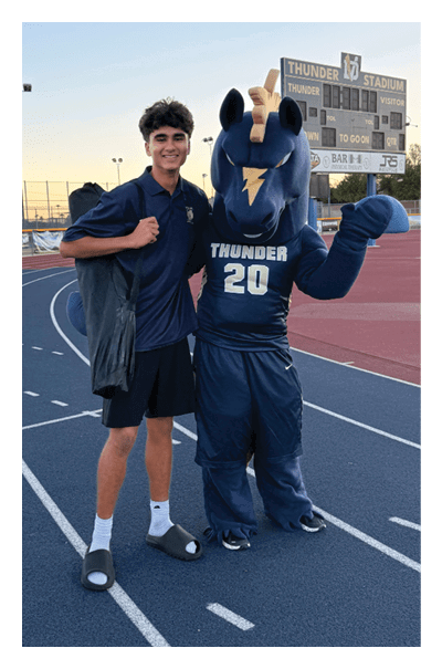 A person wearing a dark shirt and shorts stands next to a mascot dressed in a blue Thunder costume on a running track near a scoreboard at sunset. Description generated by AI
