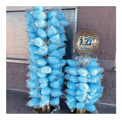 Two large balloon arrangements made of multiple small blue balloons and a central transparent balloon with a logo are displayed against a brick wall near a metal door. Description generated by AI
