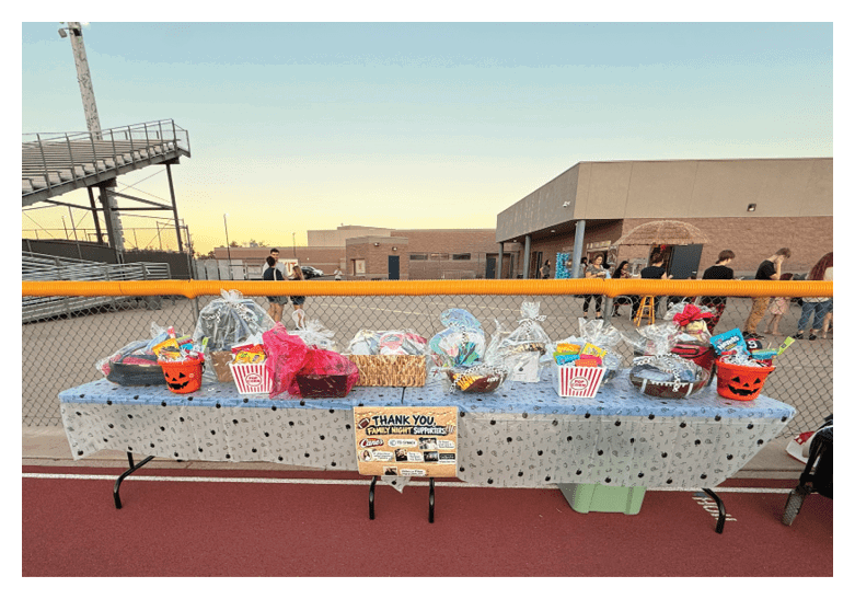 A table covered with a polka dot tablecloth displays various gift baskets and Halloween-themed containers on a running track with a school building and bleachers in the background. Description generated by AI