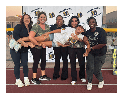 Five adults stand on a track field holding a sixth adult horizontally while smiling in front of a backdrop with logos during sunset. Description generated by AI