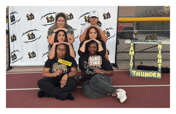 Five people are posing on a track in front of a backdrop with football logos, with two sitting on the ground and three kneeling behind them, next to a decorative frame that reads “Family Night Thunder." Description generated by AI