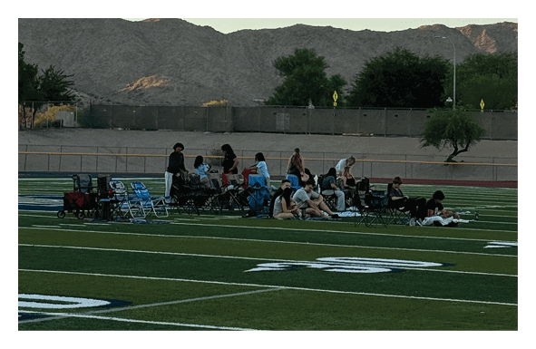People sitting and standing near the edge of a sports field with mountains and a clear sky in the background. Description generated by AI