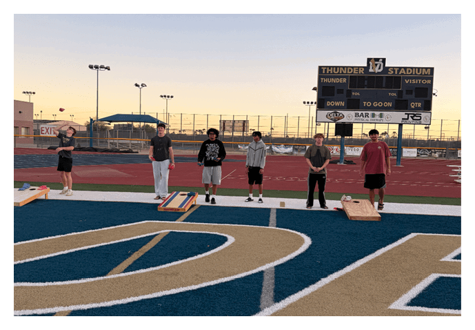 Several people stand on a track field near a scoreboard at sunset, with some holding clipboards and others wearing casual athletic clothing. Description generated by AI