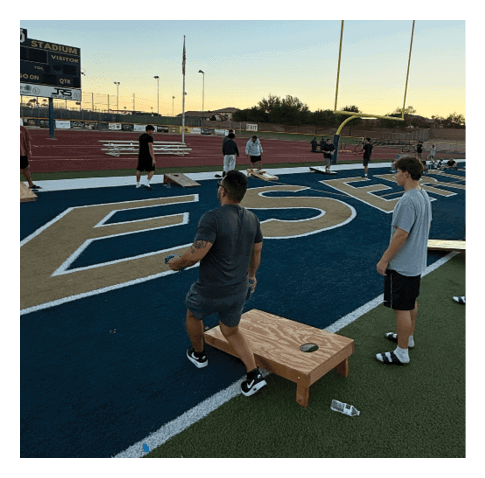Two adults stand on a sports field near a wooden cornhole board while several other people are visible in the background on a track and field area. Description generated by AI