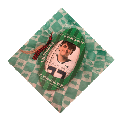 A green and white checkered gift bag with Christmas tree patterns contains a football-shaped ornament featuring a person's photo, the word “TOUCHDOWN," and a red and black plaid bow. Description generated by AI