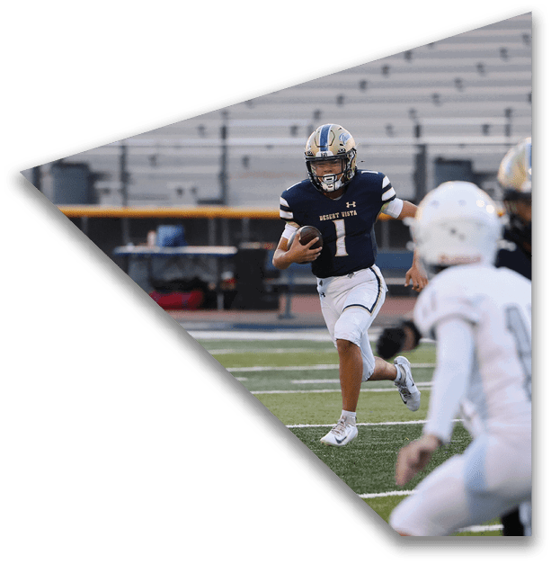 A person in a dark sports uniform is running with a football on a field while two other people in white uniforms are approaching. Description generated by AI