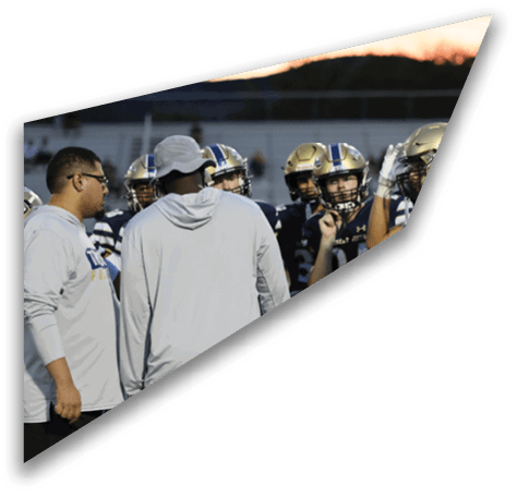 A group of people wearing football helmets and uniforms are gathered closely together on a field with two adults in casual clothing standing nearby. Description generated by AI
