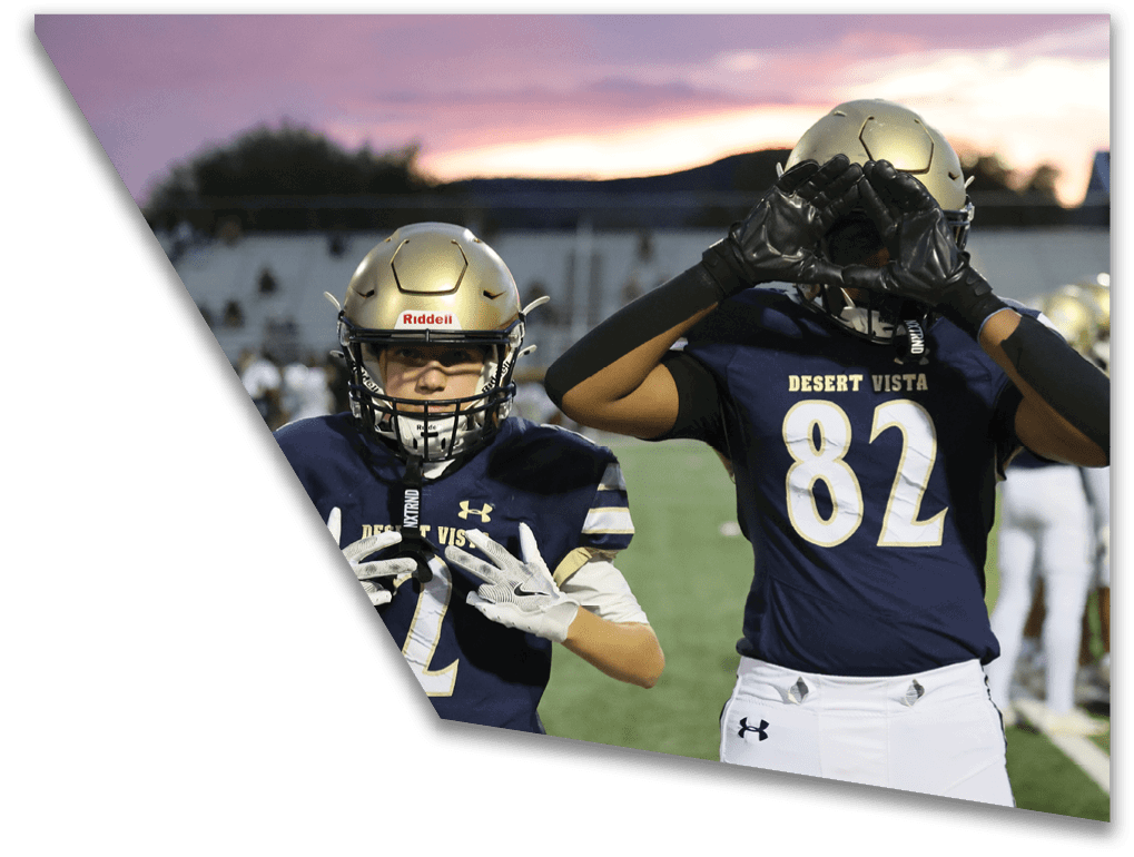 Two people wearing football uniforms and helmets stand on a field at sunset with one person holding a football near their face and the other gesturing with their hands. Description generated by AI