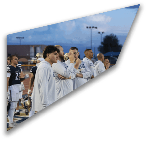 A group of people wearing white shirts stand on the sidelines of a sports field at dusk while players in football uniforms stand nearby holding helmets. Description generated by AI