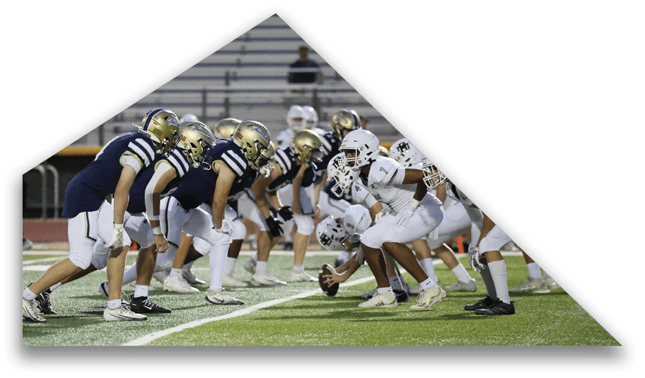 Two opposing American football teams in helmets and uniforms are lined up facing each other on a field with one player holding the ball ready to snap. Description generated by AI