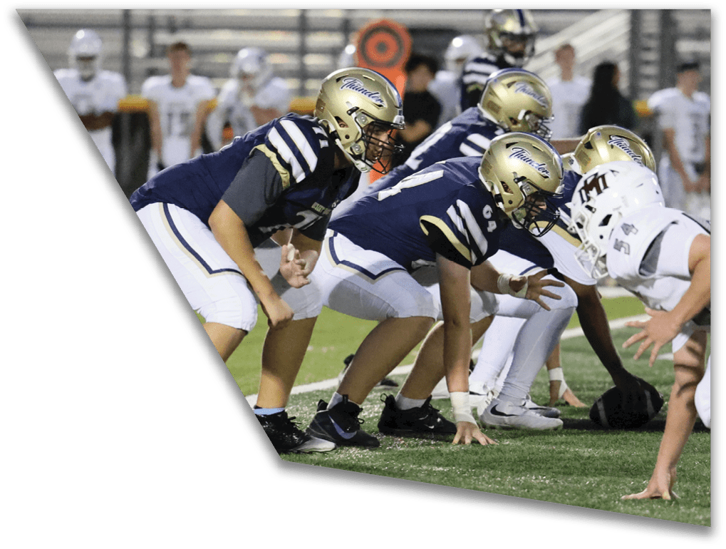 Several people in football uniforms and helmets are lined up on a field in a ready stance facing opponents in white uniforms. Description generated by AI