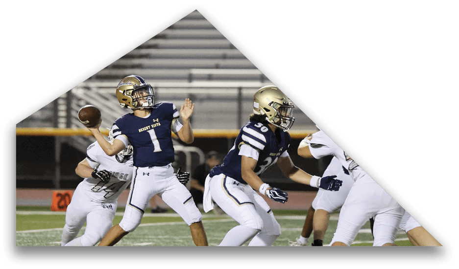 A person wearing a dark blue football uniform with the number one is preparing to throw a football while other players in similar and white uniforms are positioned around them on a field with empty bleachers in the background. Description generated by AI