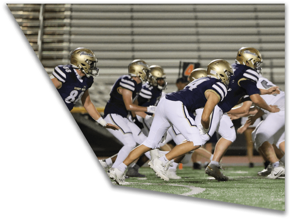 A group of people wearing helmets and sports uniforms are lined up in a crouched position on a football field, appearing ready to start a play. Description generated by AI