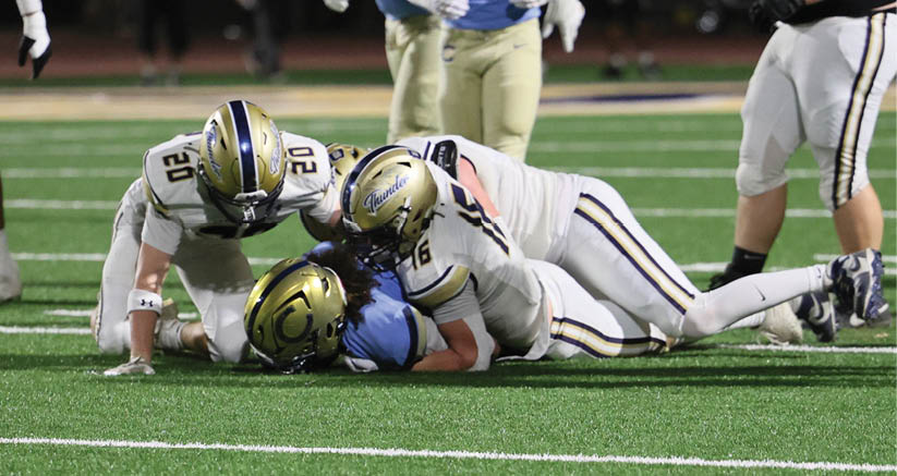 Three people in football uniforms are on the ground during a game on a turf field with one player tackling another while a third player is nearby. Description generated by AI