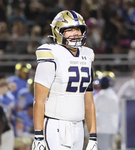 A person wearing a football helmet and a white jersey with the number 22 and the words Desert Vista is standing on a field with a blurred crowd in the background. Description generated by AI