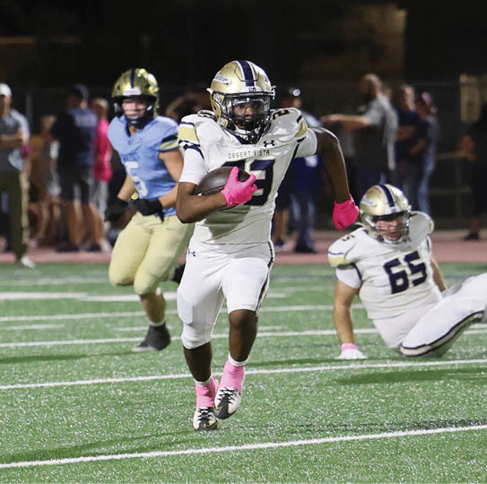 A person in a white football uniform with pink gloves and socks is running with the ball on a field while another person in a white uniform is on the ground and a person in a blue uniform is chasing behind. Description generated by AI