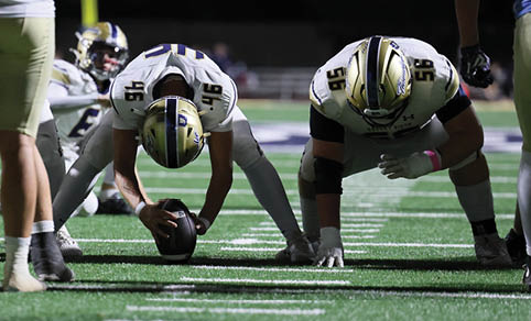 Two people in football uniforms are bent over on a field near a goalpost, with others standing or kneeling nearby during a nighttime game. Description generated by AI