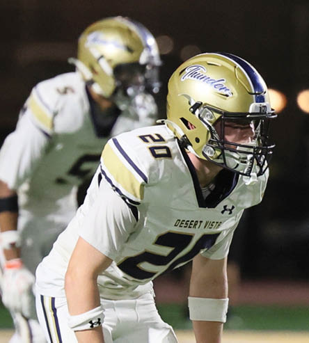 A person wearing a white football uniform with the number 20 and a helmet is leaning forward in a ready stance on a field. Description generated by AI