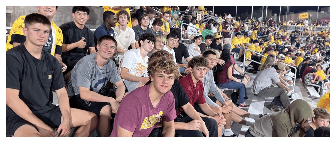 A large group of people wearing casual clothing are seated closely together on bleachers at an outdoor nighttime event, with some smiling and interacting. Description generated by AI