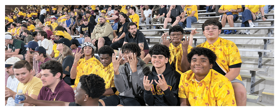 A group of people wearing yellow shirts and casual clothing are sitting on bleachers at an indoor event with some playing brass instruments in the background. Description generated by AI