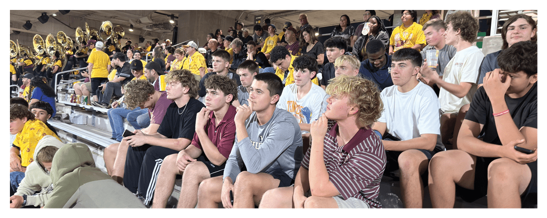 A large group of people wearing casual clothing and some in yellow shirts are seated closely together on bleachers in an indoor setting, attentively watching an event. Description generated by AI