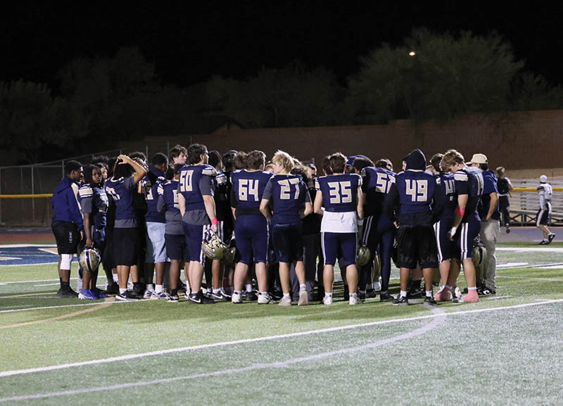 A group of people wearing dark sports uniforms are huddled together on a football field at night while other players stand in the background. Description generated by AI