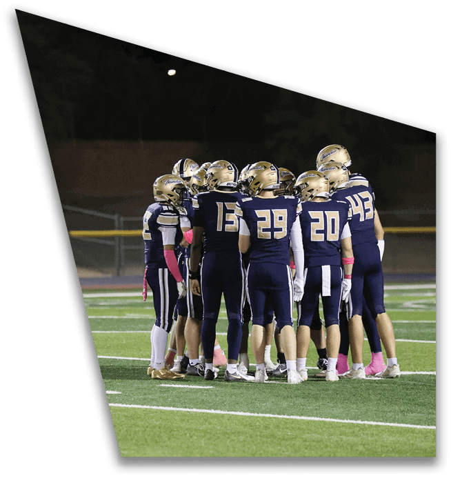 A group of football players wearing dark uniforms and gold helmets are huddled together on a green field at night. Description generated by AI